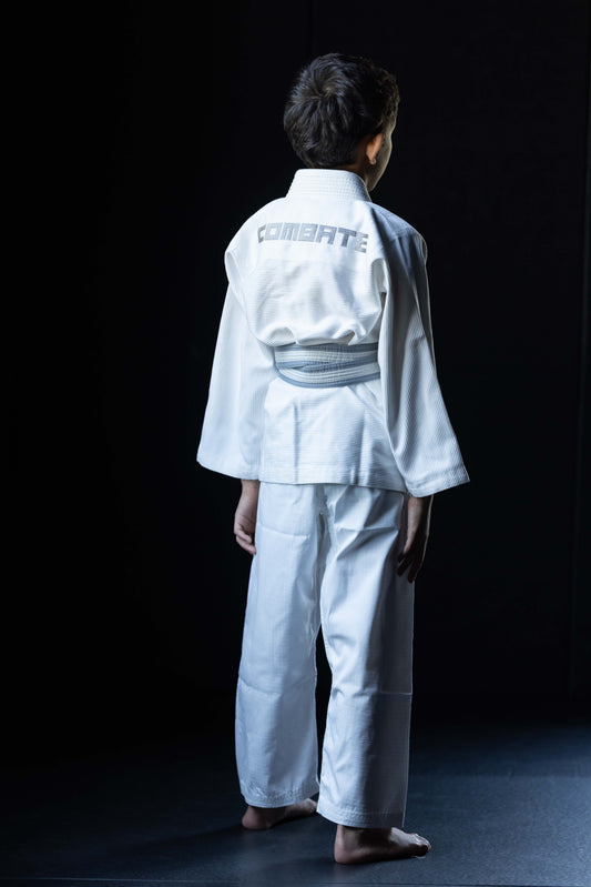 A child wearing a white martial arts gi with a gray belt, shown from the back against a dark studio background. The word 'COMBATE' is printed on the back of the collar.
