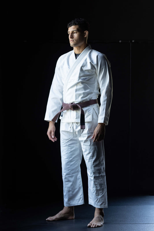 A man wearing a white Brazilian Jiu-Jitsu (BJJ) gi with a brown belt, standing in a studio with a black background.
