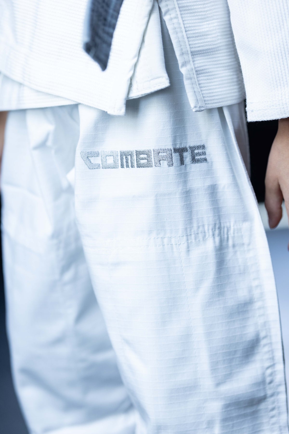Close-up of a white martial arts gi with the word 'COMBATE' printed in gray on the thigh, worn by a person whose hand is partially visible.