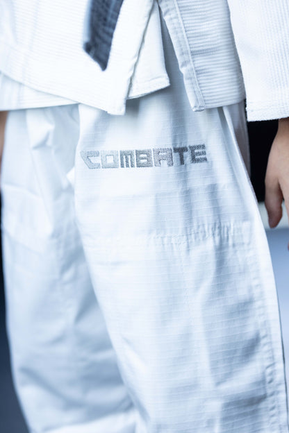 Close-up of a white martial arts gi with the word 'COMBATE' printed in gray on the thigh, worn by a person whose hand is partially visible.