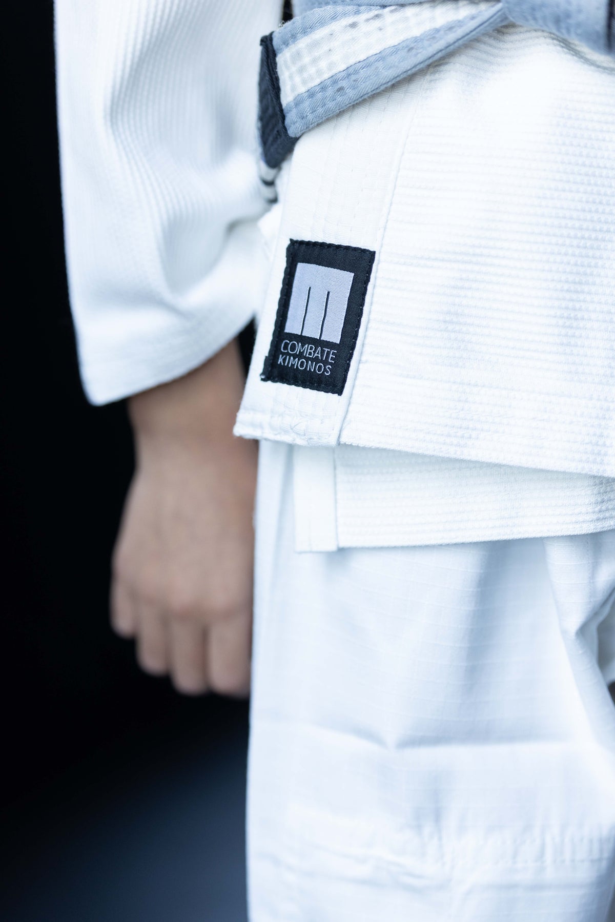 Close-up of a white martial arts gi with a black and gray belt, featuring a black brand patch that reads 'COMBATE Kimonos'.