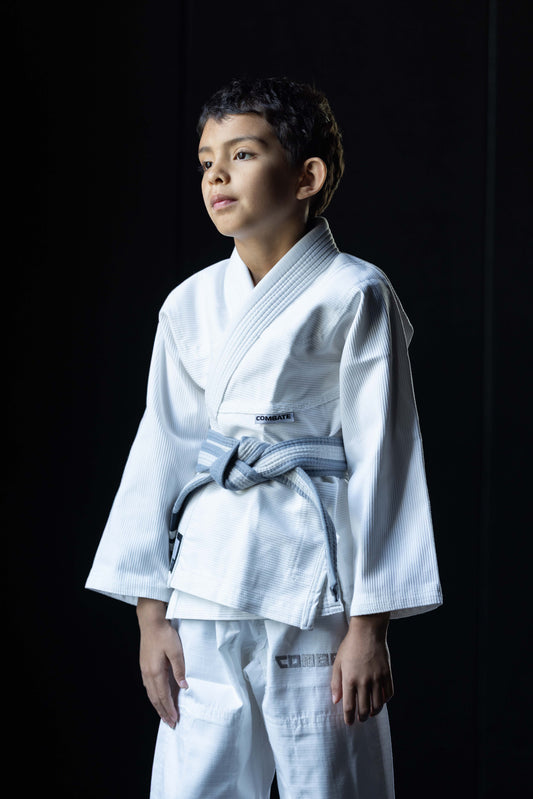 A young boy wearing a white martial arts gi with a greywhite belt, standing against a dark background.