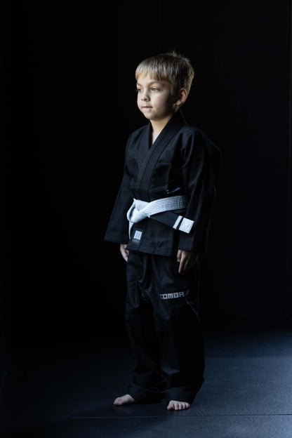 A young boy wearing a black martial arts gi with a white belt, standing in a studio against a dark background.