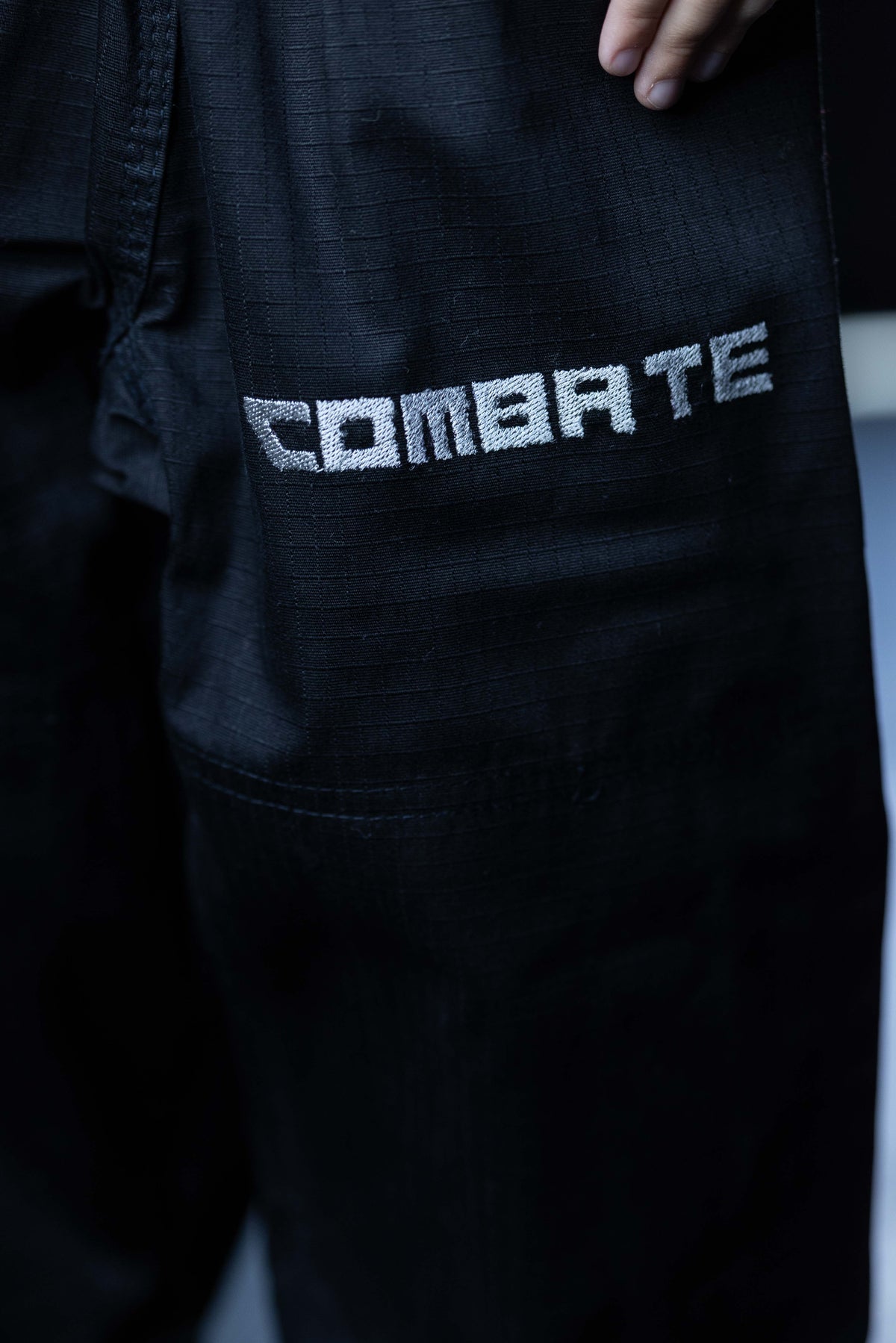 Close-up of black martial arts gi pants featuring a silver textured 'COMBATE' logo on the thigh, with a hand visible at the top edge.