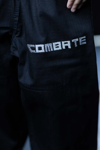 Close-up of black martial arts gi pants featuring a silver textured 'COMBATE' logo on the thigh, with a hand visible at the top edge.